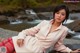 A woman in a pink coat sitting on a rock by a river.