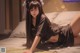 A woman in a black satin outfit reclines on a bed with pillows.
